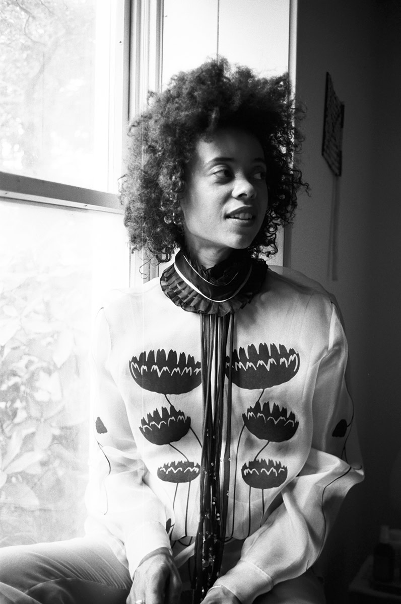 Elegant black and white portrait of a woman with natural curly hair, wearing a floral-patterned blouse, sitting by a window. Captures artistic portrait photography by Pamela Hanson, emphasizing natural beauty and sophisticated style.