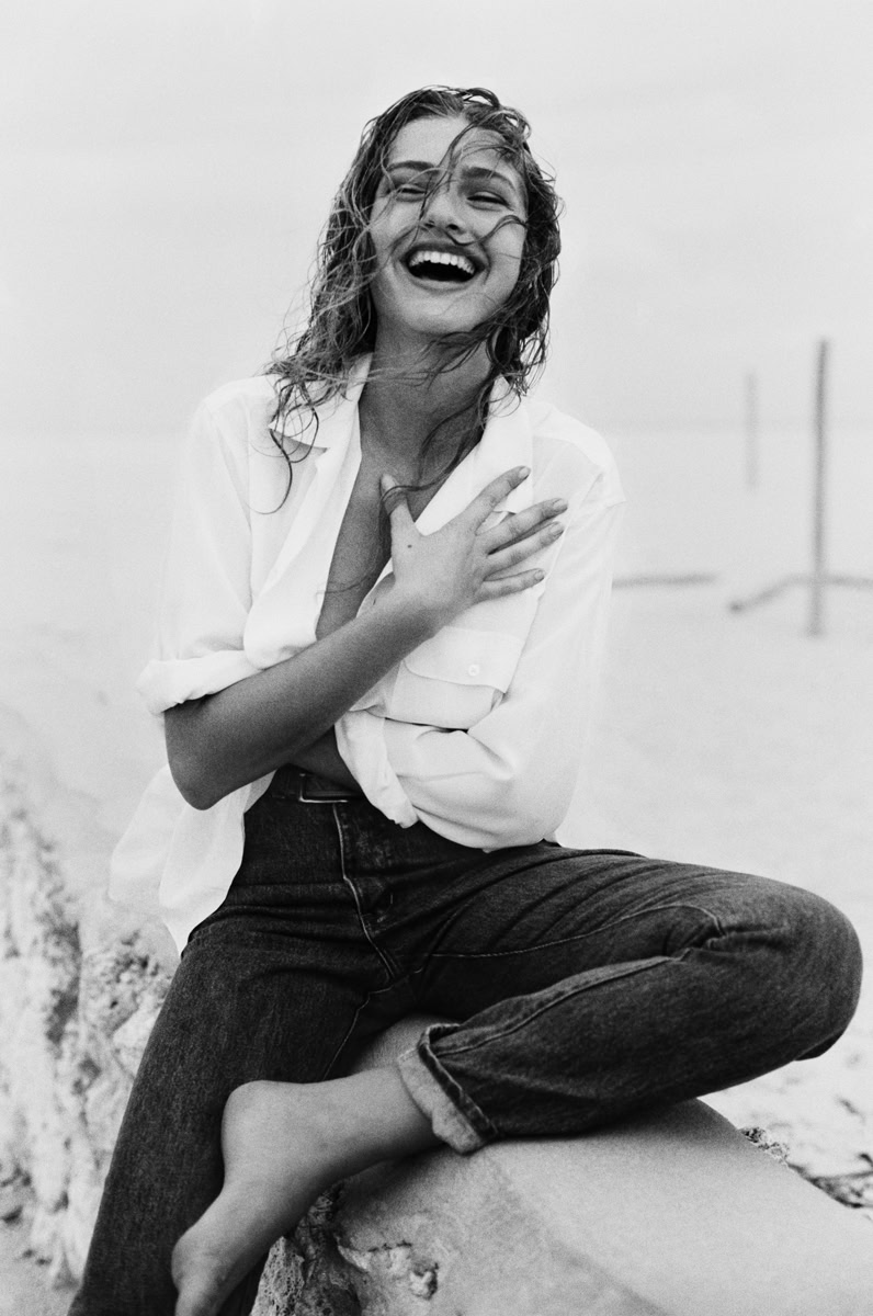 Stunning black and white portrait of a joyful woman with wet hair, sitting on a beach rock, embodying natural beauty, candid happiness, and professional fashion photography by Pamela Hanson.