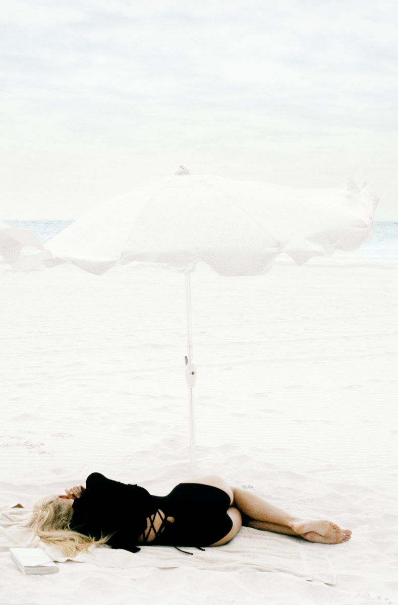 Soft-focus beach photography by Pamela Hanson, featuring a woman under a white parasol on sandy shore. Elegant, artistic portrait emphasizing beach lifestyle, fashion, and relaxation.