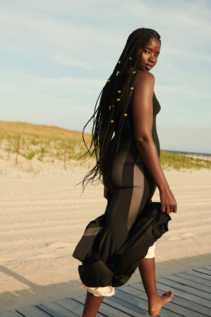 Elegant beach portrait of a woman with long braided hair, dressed in stylish black and beige flowing fashion, standing on a wooden boardwalk at sunset with a sandy beach and dunes in the background.