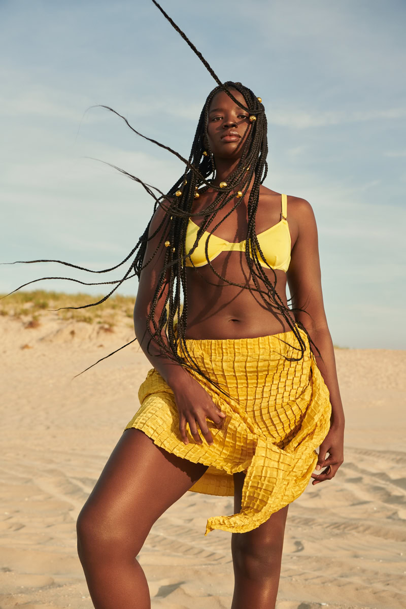 Vibrant woman in yellow swimsuit and skirt posing on beach with breezy hair, natural sunlight, and sandy dunes in the background, showcasing stunning beach fashion photography by Pamela Hanson.