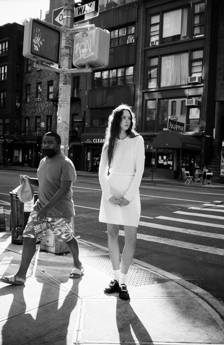 Urban street scene featuring a young woman in a white dress and loafers, with a man in casual clothing walking nearby, captured in black and white photography by Pamela Hanson.