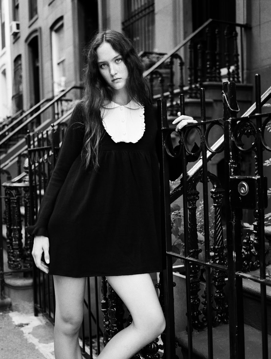 Black and white portrait of a young woman with long wavy hair, wearing a vintage-style black dress with a white collar, standing next to an ornate iron fence in an urban setting, showcasing fine art fashion photography.