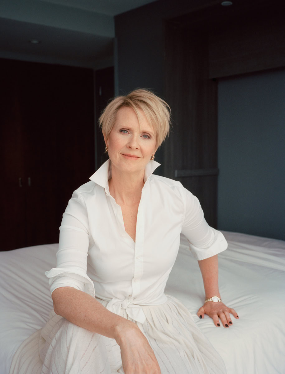 Elegant mature woman portrait in a stylish white blouse and skirt, sitting on a bed in a modern hotel room, captured by Pamela Hanson Photography.