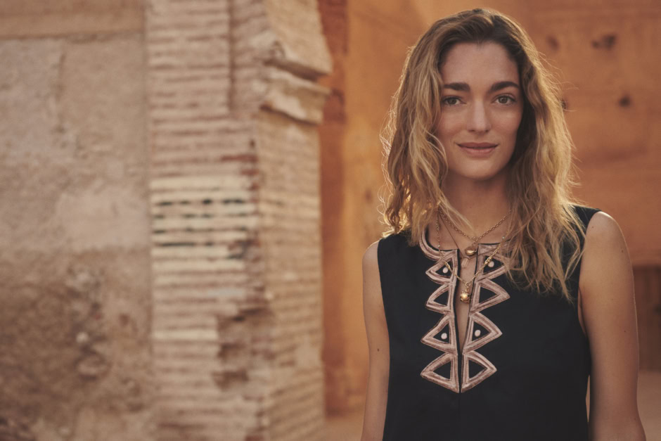 Elegant woman with wavy blonde hair wearing a stylish black embroidered dress, accessorized with layered gold necklaces, posing indoors with warm wooden and brick background for fashion photography.