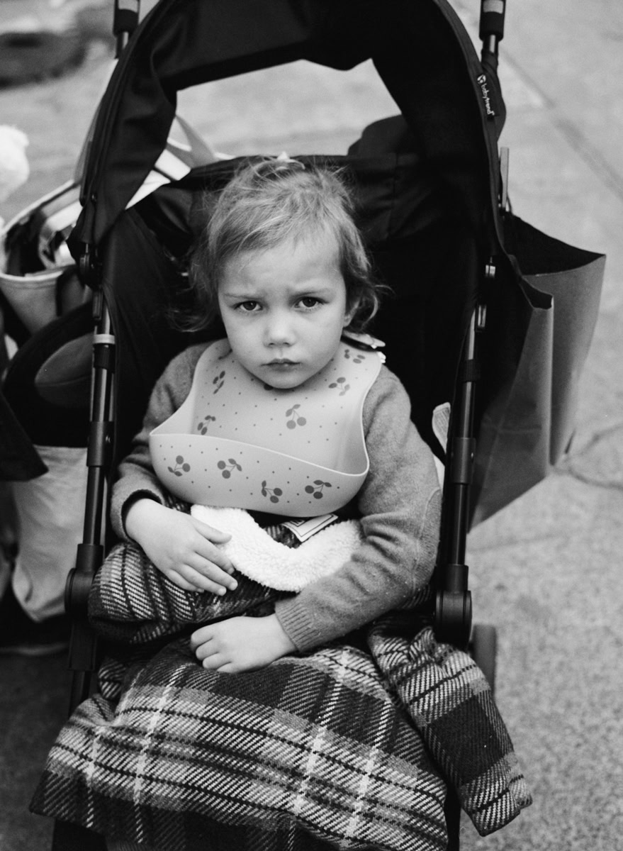 A young girl with a serious expression sitting in a stroller, black and white photography, capturing candid childhood moments, emotional portrait, street photography, Pamela Hanson photography.