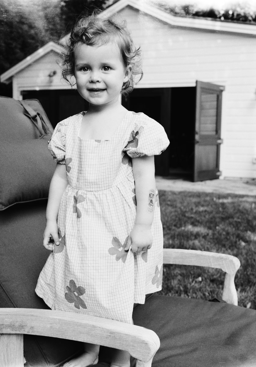 Cute young girl with curly hair standing outdoors in front of a shed, black and white portrait.