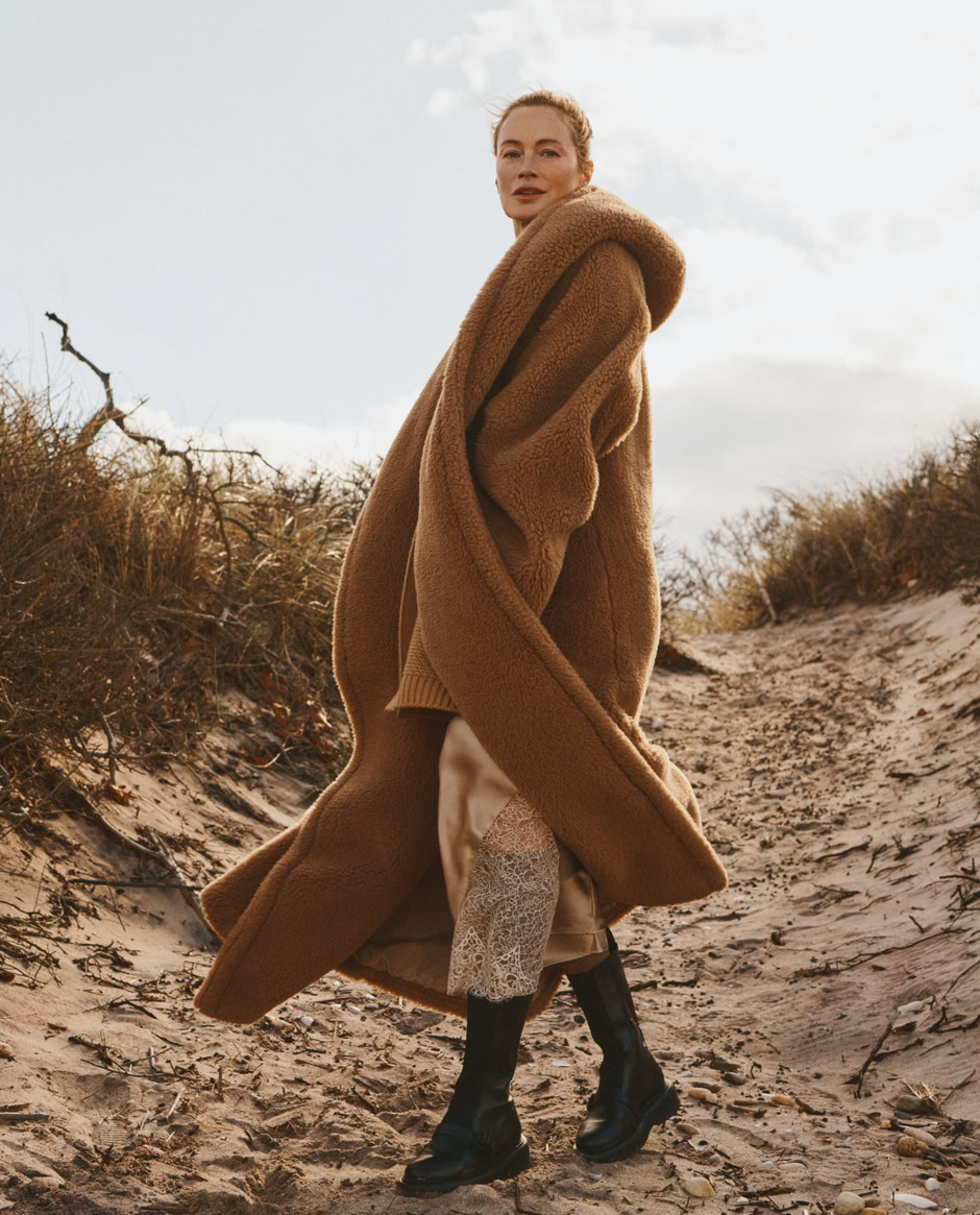 Warm winter fashion shot featuring a woman in a cozy teddy coat, layered with patterned dress and chunky black boots, captured on a sandy trail with dry bushes under a cloudy sky.