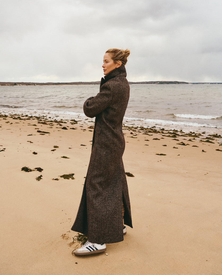 Stylish woman in a long herringbone coat standing on beach with waves and cloudy sky, modern fashion photography by Pamela Hanson.