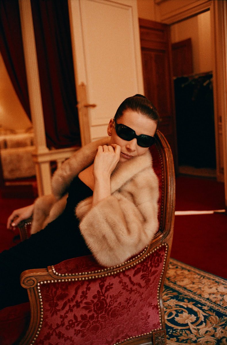 Elegant woman in luxury fur coat and dark sunglasses sitting on ornate vintage armchair in sophisticated interior.