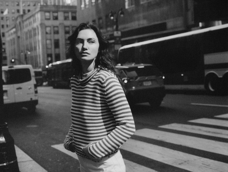 A woman walking on a busy city street in black and white, wearing a striped sweater and looking confidently into the camera, capturing urban fashion and street style photography.