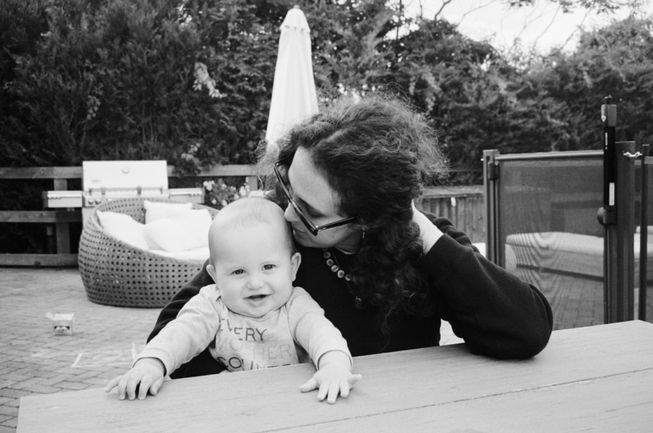 A black and white photograph of a woman with curly hair and glasses gently kissing her smiling baby on the head outdoors, showcasing intimate family moments captured by Pamela Hanson Photography.