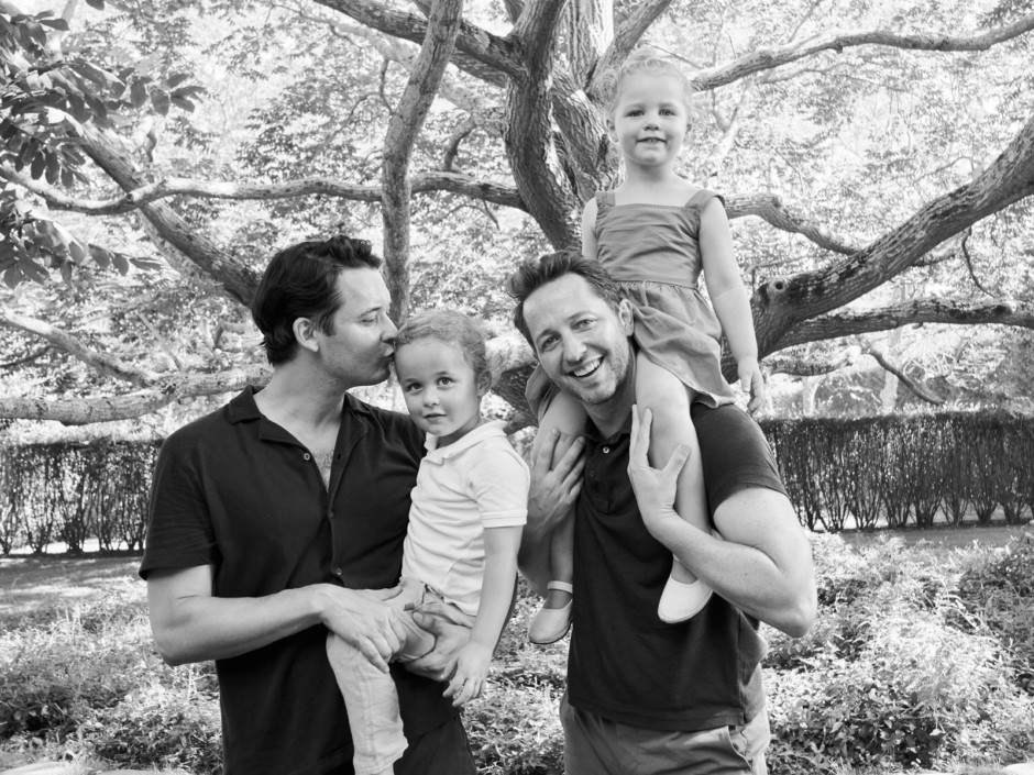 Family outdoor park portrait photography featuring two dads and two young daughters, black and white image capturing joyful candid moments with trees in the background.
