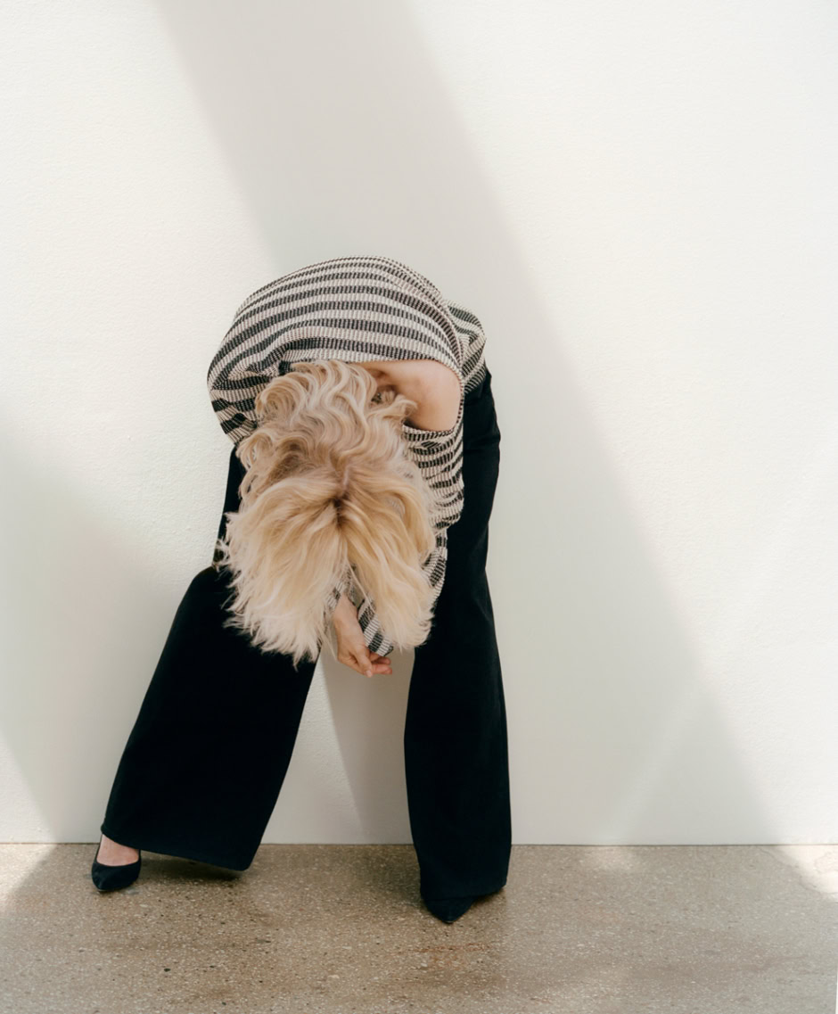 Elegant woman with blonde wavy hair, wearing wide-leg black trousers and a striped top, bending down to pet a fluffy golden retriever, in a minimalistic white room, captured by Pamela Hanson Photography.