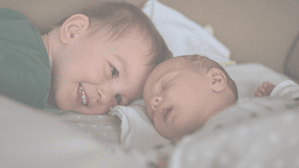 A smiling toddler boy lying beside his sleeping newborn sibling