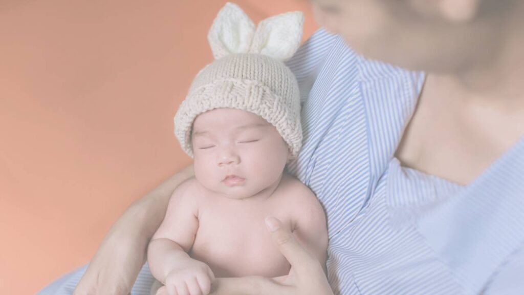 A sleeping newborn in a knitted bunny ear bonnet held against a parent's chest