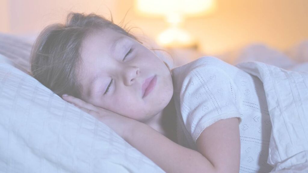 A young girl asleep on a blue pillow with a lamp glowing behind her