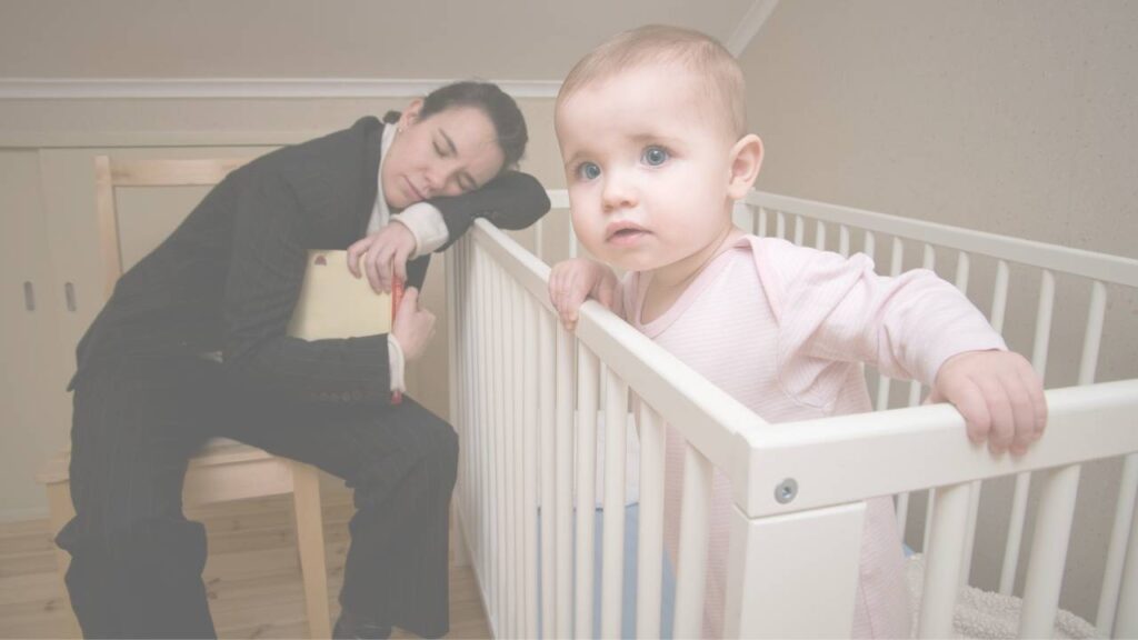 An exhausted parent asleep on a chair beside a cot while the baby stands up wide awake