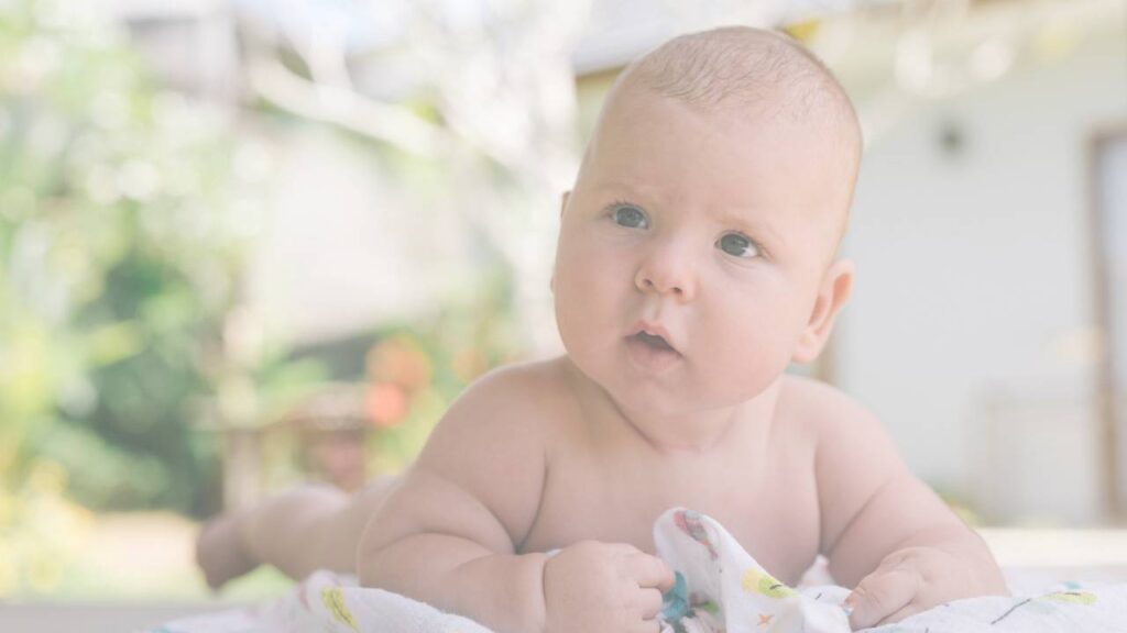 A baby lying on their tummy outdoors on a blanket, looking at the camera
