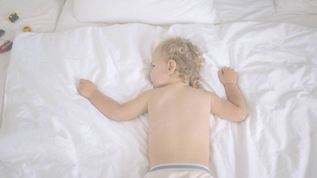A curly haired toddler asleep face down across a big white bed