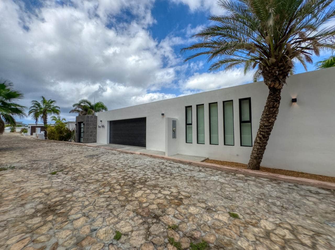 Modern White Pedregal House With Large Vertical Windows And A Black Garage Door With Palm Trees And A Cobblestone Driveway.