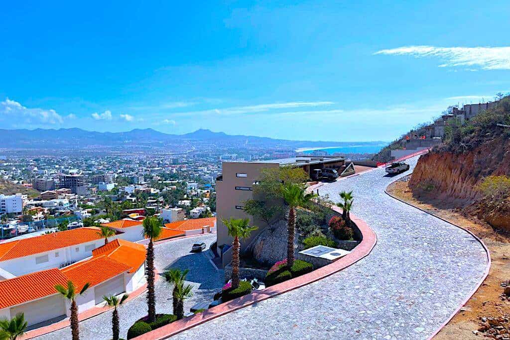 Pedregal Hillside Road With Ocean Views, Showcasing A Winding Cobblestone Street Lined With Palm Trees And Modern Homes, Offering Stunning Vistas Of The Pacific Ocean.