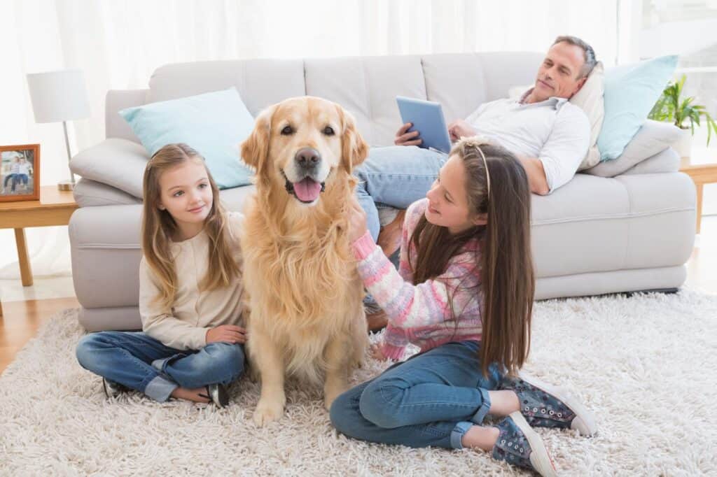 Two girls sit on a carpet petting a golden retriever, while a man reclines on a sofa in the background, tablet in hand, reminding himself to schedule AC service.