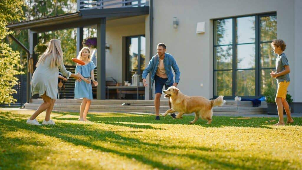 The family enjoys a game of frisbee on the grassy lawn beside their modern house, where an efficient HVAC system keeps seasonal allergies at bay. Under a clear sky, two adults, two children, and a golden retriever revel in carefree play.