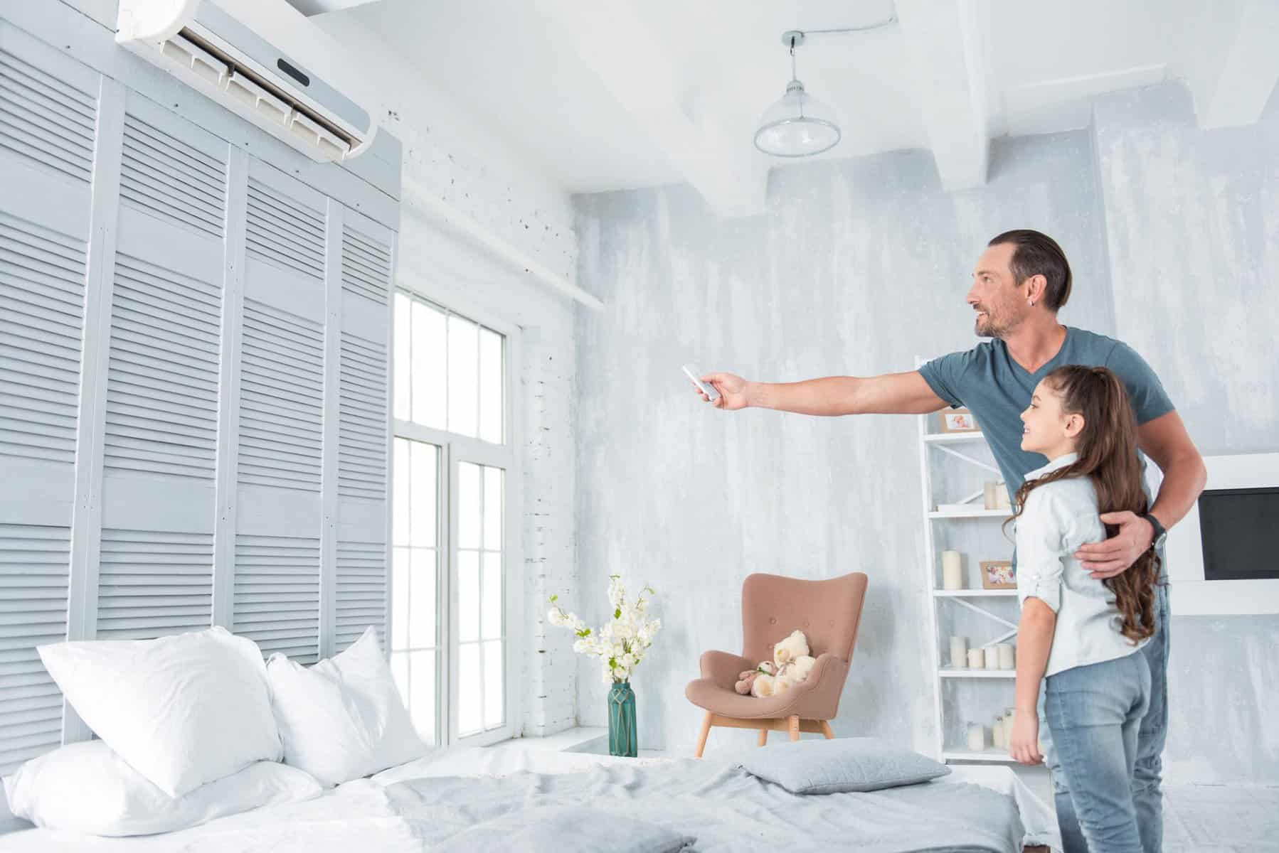 A man cradles a child in his arms as he points a remote at the mini split in their bright bedroom, elegantly adorned with white and gray decor.