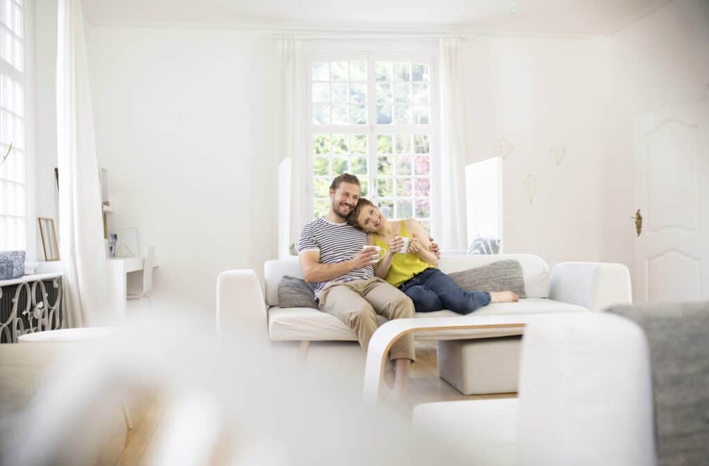 A man and a woman sit on a white couch in a bright room with a large window, sipping drinks as they chat about their latest DIY heating system maintenance adventure.
