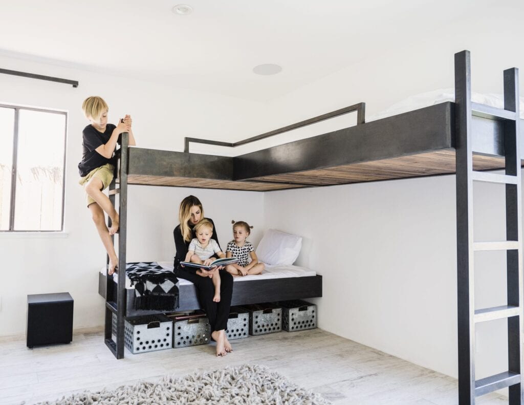 A woman sits with two young children on a lower bed under a lofted bed. A child climbs a ladder to the top bunk in their bright, minimalist room, designed with home ventilation in mind for healthier indoor air.