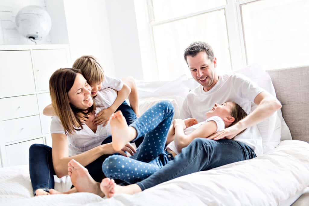 A family of four laughs and plays on a bed in a bright, airy room. The parents and two kids, all in casual white tops and blue jeans, enjoy the warm air gently flowing from the AC.