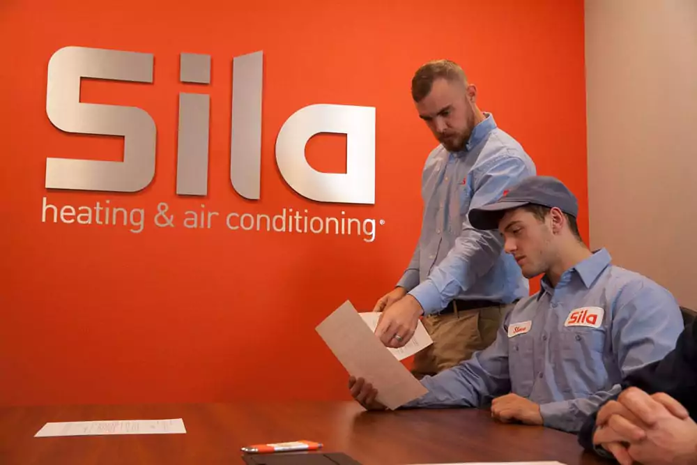 Two workers in Sila uniforms discuss an "About Us" document in an office, framed by an orange wall adorned with the Sila Heating & Air Conditioning logo.