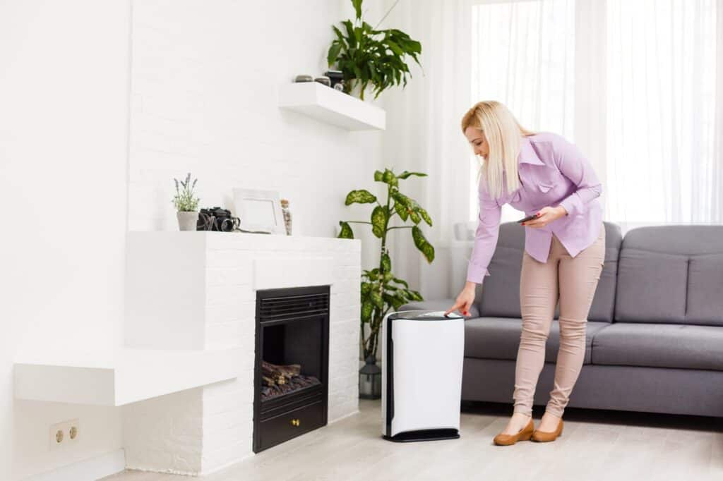 In a bright living room adorned with plants and a gray couch, a woman adjusts her white air purifier—one of the easy ways to control home humidity and maintain comfort.