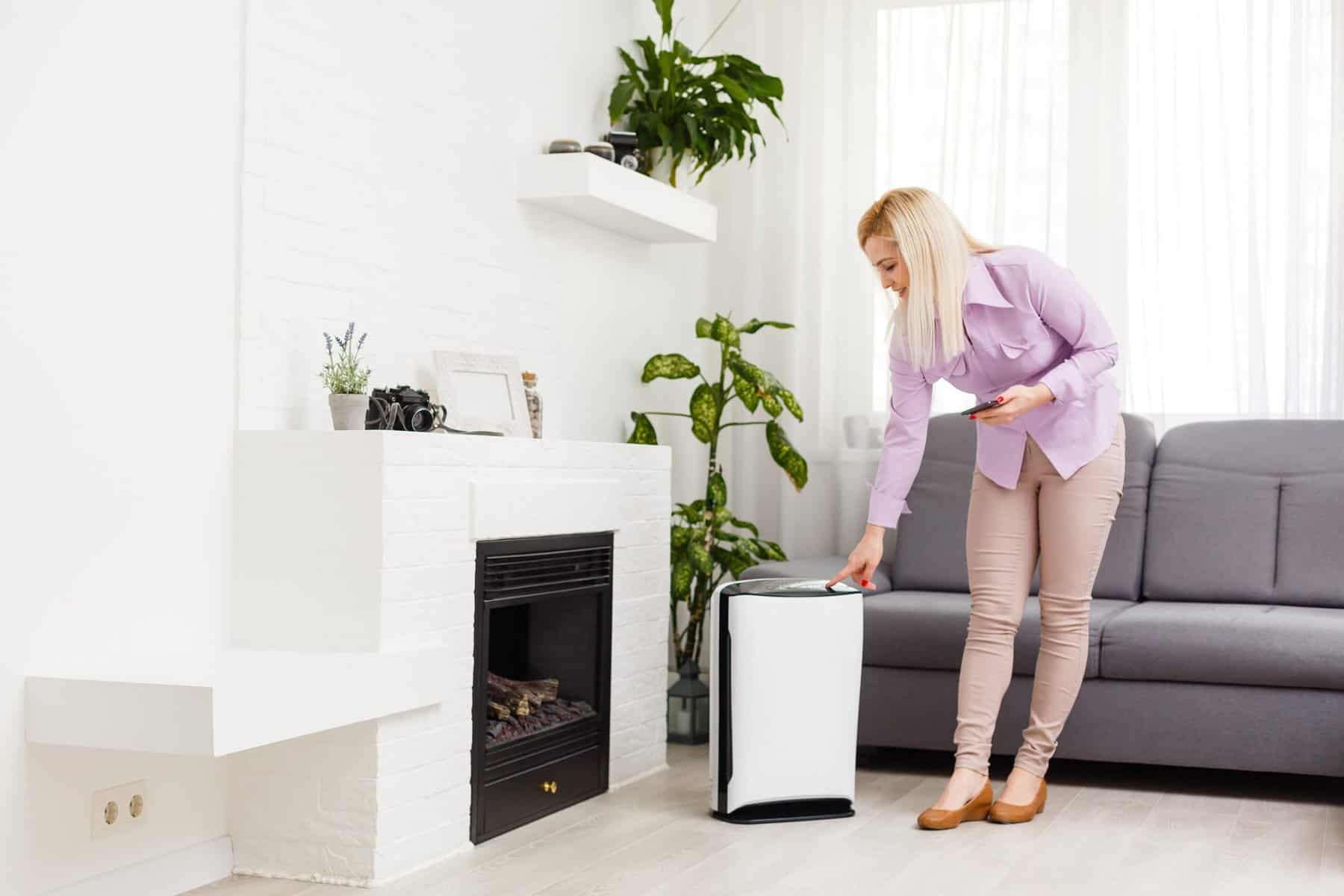 In a bright living room adorned with plants and a gray couch, a woman adjusts her white air purifier—one of the easy ways to control home humidity and maintain comfort.