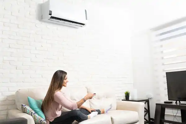 In her cozy Philadelphia apartment, a woman relaxes on the sofa with a remote control, facing the television and an air conditioner on the white brick wall, ensuring she schedules regular mini-split maintenance to keep her PA home comfortable year-round.