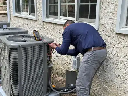 A technician is inspecting and repairing an outdoor air conditioning unit next to a house, using a digital multimeter to check the system.