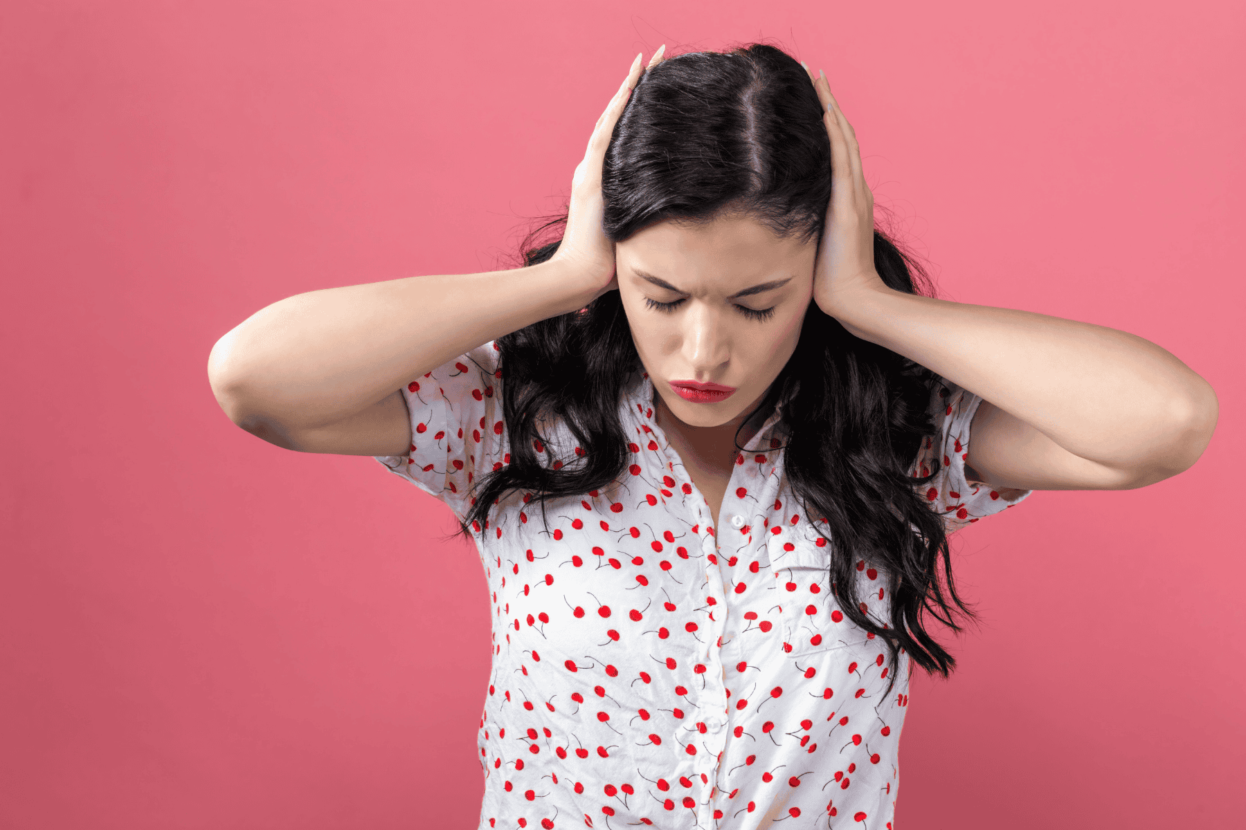 Woman with long dark hair covers her ears with both hands, wearing a white shirt with red cherry prints, standing against a solid pink background—her expression clearly showing the frustration familiar to anyone dealing with banging pipes or the irritating water hammer often found in home plumbing.