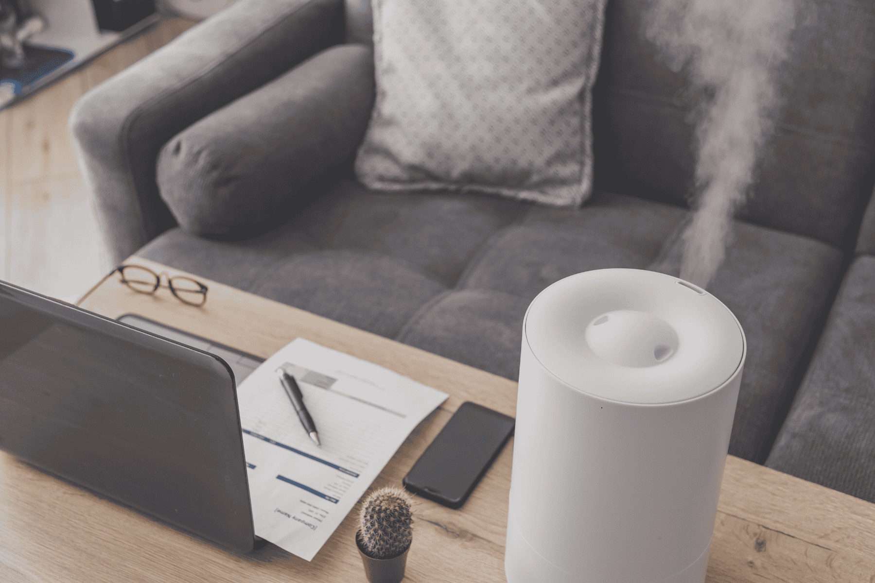 A white humidifier emitting mist sits on a table next to a laptop, papers, glasses, a smartphone, and a small cactus with a gray couch in the background—perfect for combating dry air in the house and maintaining optimal indoor humidity during winter.