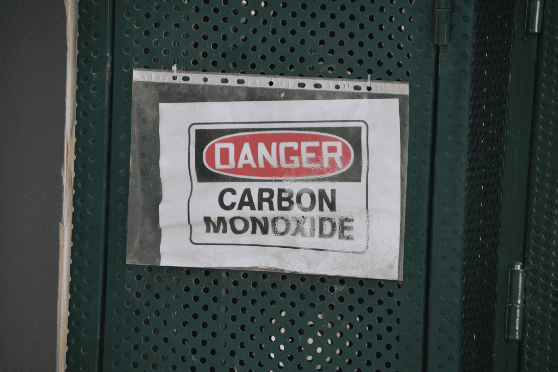 A sign on a perforated green metal wall reads “DANGER CARBON MONOXIDE” in black and red text, following safety guidelines for proper placement of carbon monoxide detector warnings.