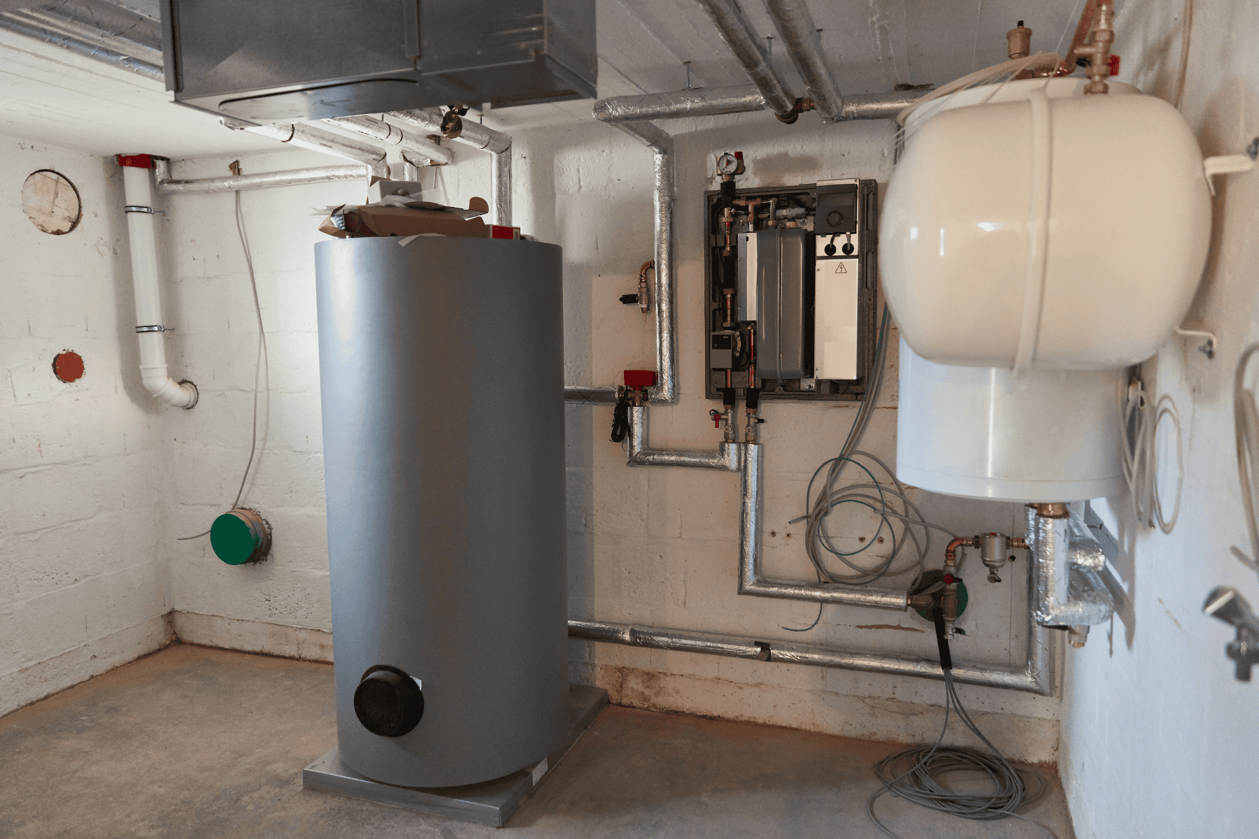 Boiler room with a large grey water heater tank, white expansion vessel, exposed pipes, and control panel—ideal for monitoring boiler warning signs or addressing boiler pressure low issues.