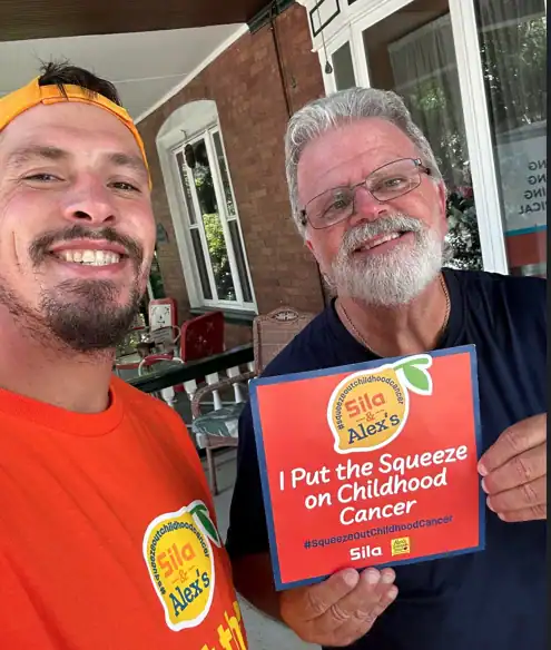 Two men smile at the camera; one holds a sign reading “I Put the Squeeze on Childhood Cancer.” Both wear orange shirts proudly displaying the Alex’s Lemonade logo.
