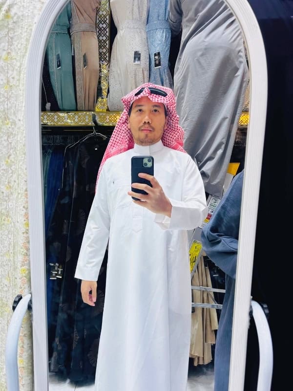 Traveler trying on a traditional white Saudi thobe and red-and-white checkered keffiyeh while taking a mirror selfie in a clothing store.