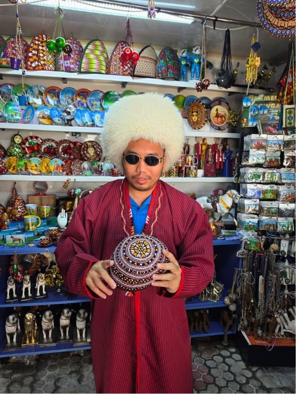 Traveler wearing a traditional burgundy Turkmen robe and large white fur hat while holding an embroidered traditional hat in a souvenir shop filled with colorful Central Asian crafts.