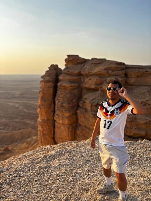 Traveler wearing a Germany football jersey and sunglasses while exploring the dramatic sandstone cliffs of Saudi Arabia at sunset.