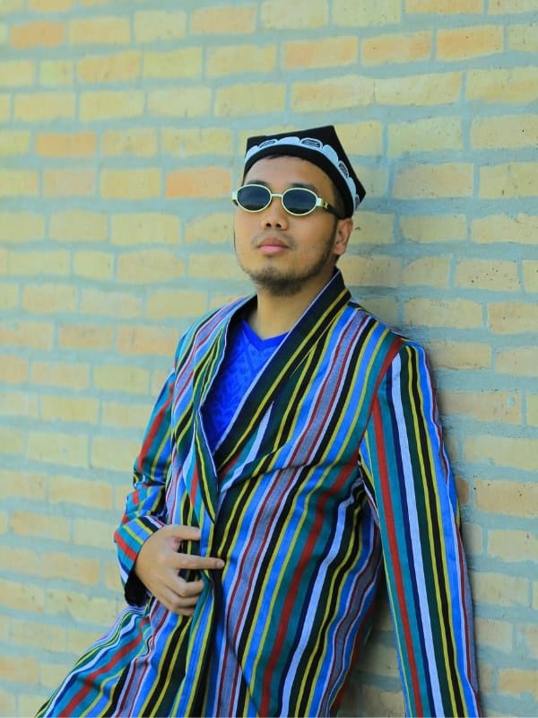 Traveler wearing a colorful traditional Uzbek striped robe, embroidered skullcap, and round sunglasses while posing against a brick wall.
