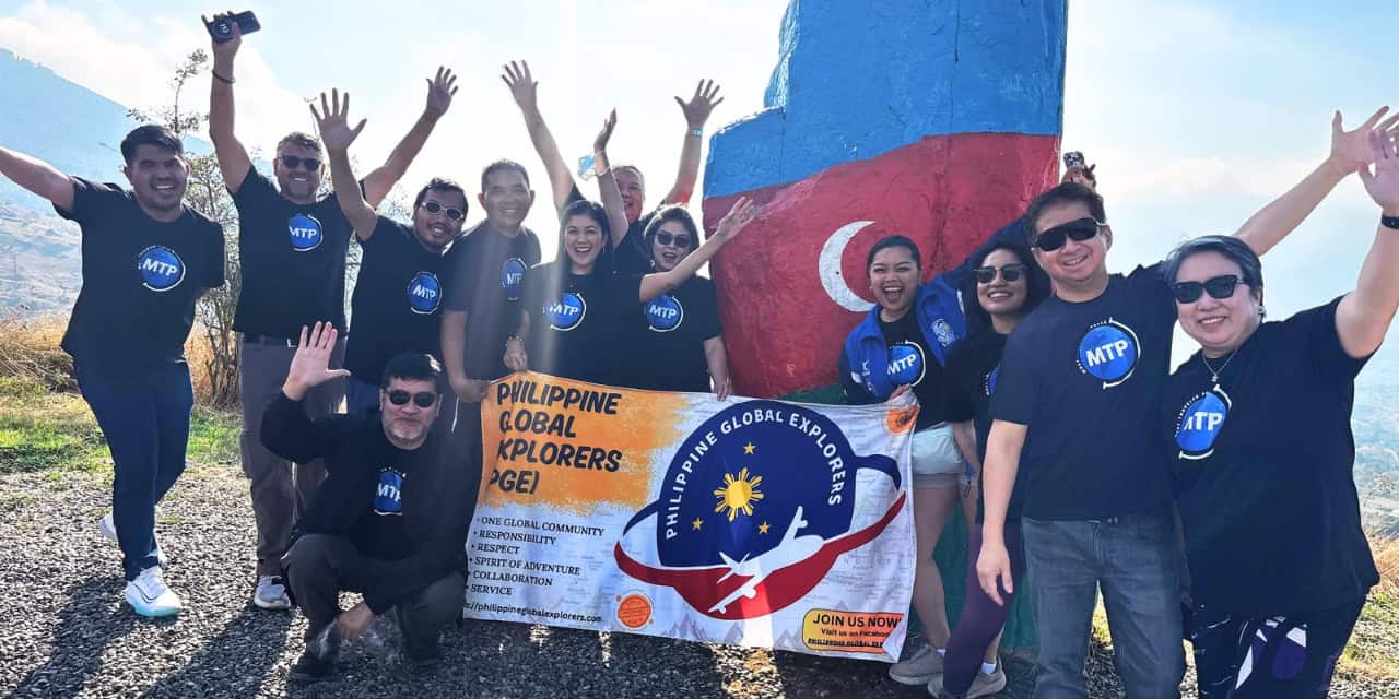Members of the Philippine Global Explorers posing with their group banner and raising their hands beside a large Azerbaijan flag monument during a group trip.