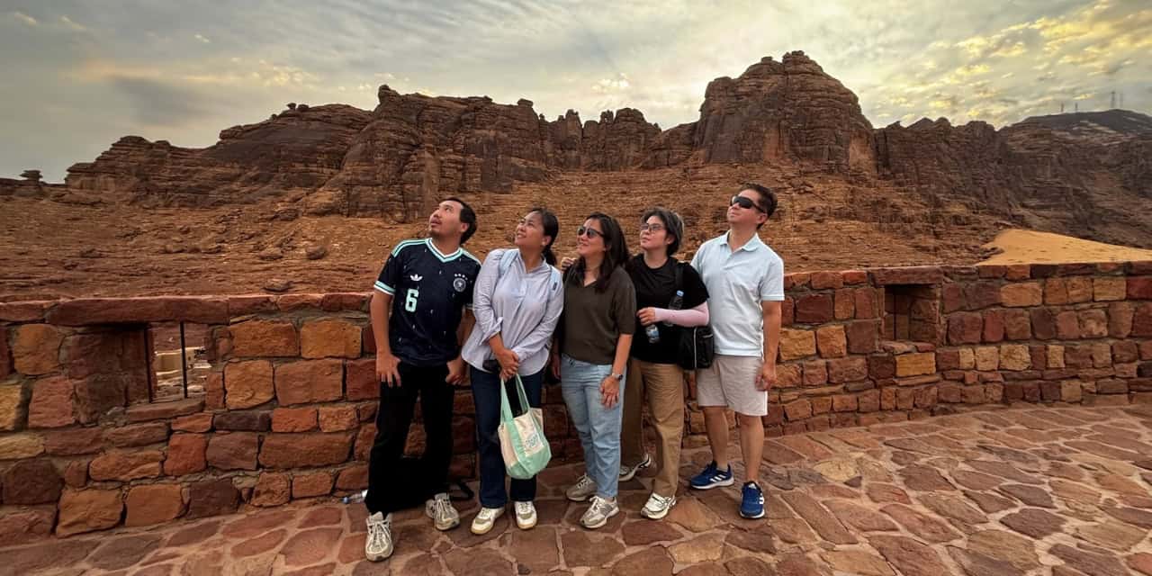 Five travelers posing together on a stone terrace overlooking the dramatic red sandstone mountains of AlUla, Saudi Arabia.