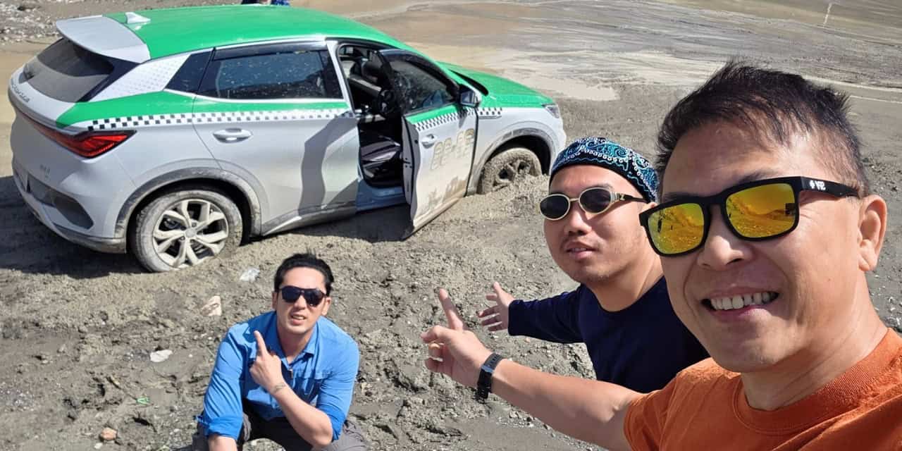 Three travelers posing for a selfie beside a green and white taxi on a rugged, rocky landscape in Uzbekistan.