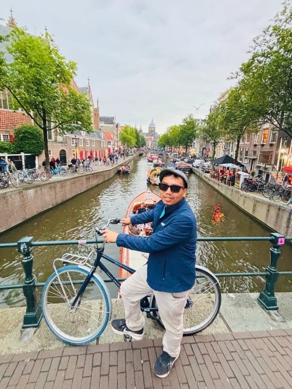 Traveler posing with a bicycle on a bridge overlooking a scenic canal lined with historic buildings and boats.